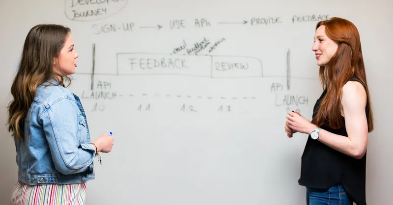 Two women engaging in a discussion about API development processes at a whiteboard.
