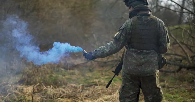 A soldier in camouflage uses a blue smoke signal during a tactical exercise in a forest setting.