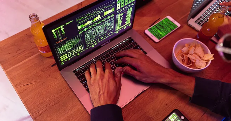 Hands typing on a laptop with coding, phone on desk, symbolizing cybersecurity.