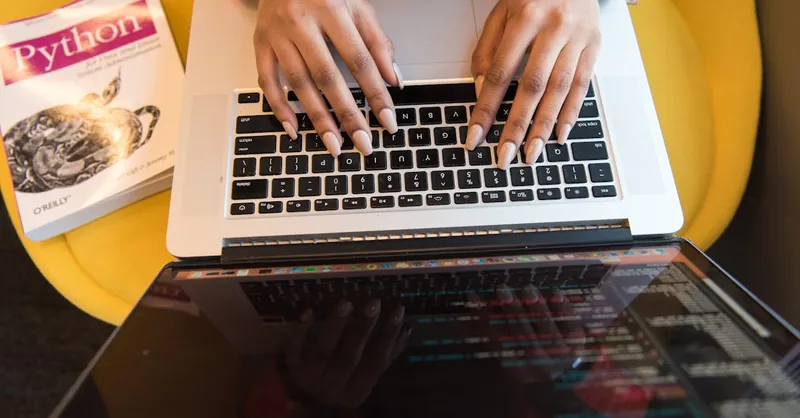 Close-up of hands typing on a laptop keyboard, Python book in sight, coding in progress.