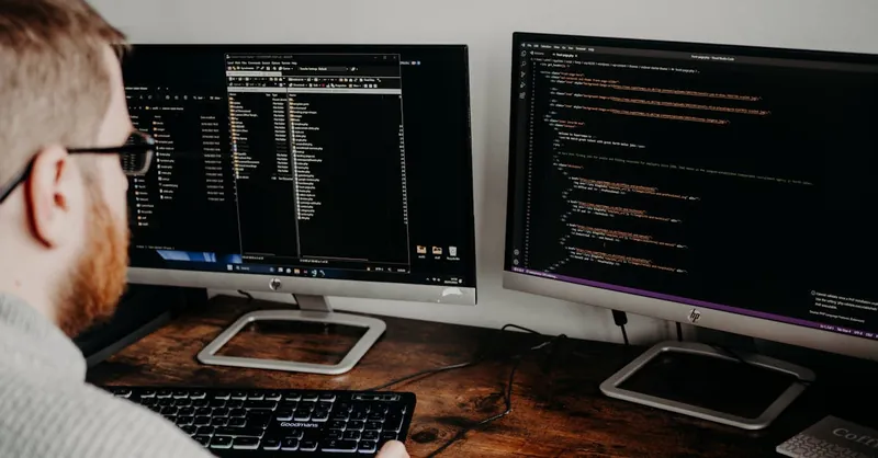 Software developer working on code with dual monitors in a home office setting.