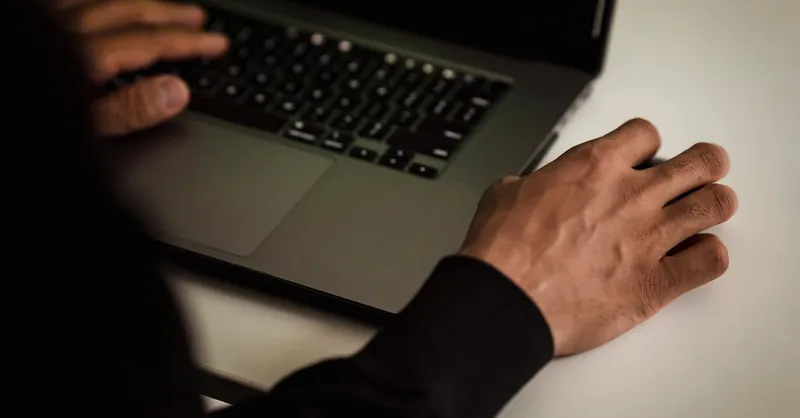 From above crop faceless male developer in black hoodie writing software code on netbook while working in light studio